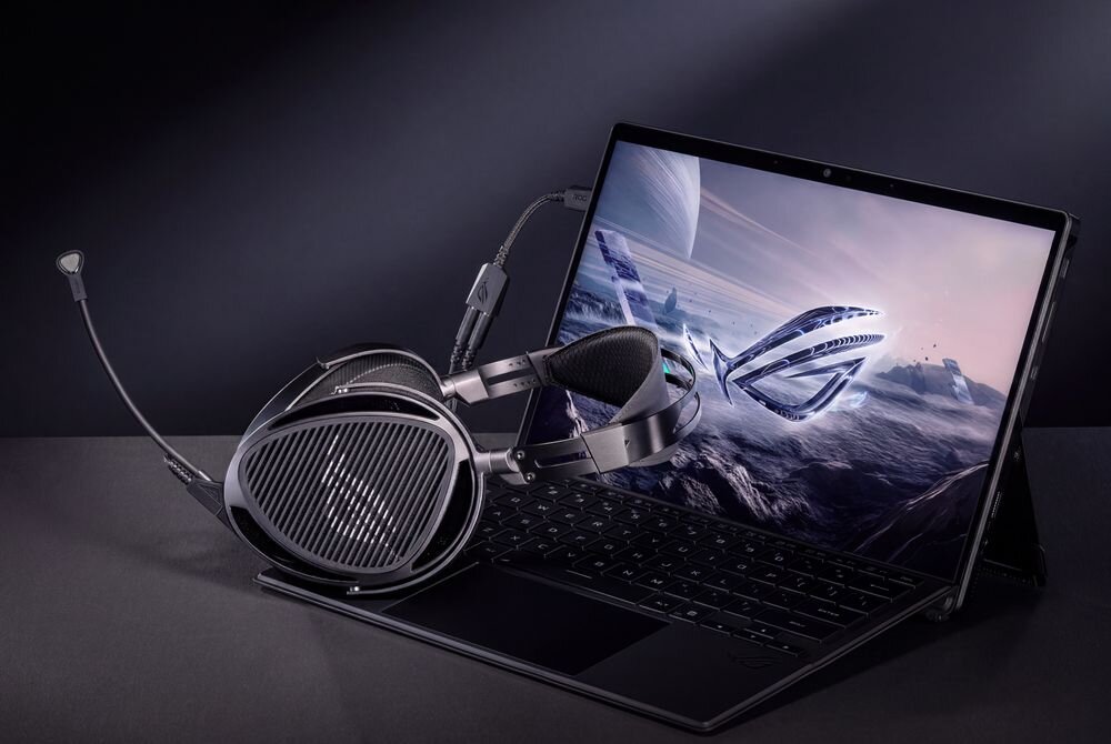 Na obrazie widać laptop z wyświetlonym futurystycznym krajobrazem oraz słuchawki nauszne umieszczone na klawiaturze. Logo ASUS jest widoczne na ekranie laptopa.