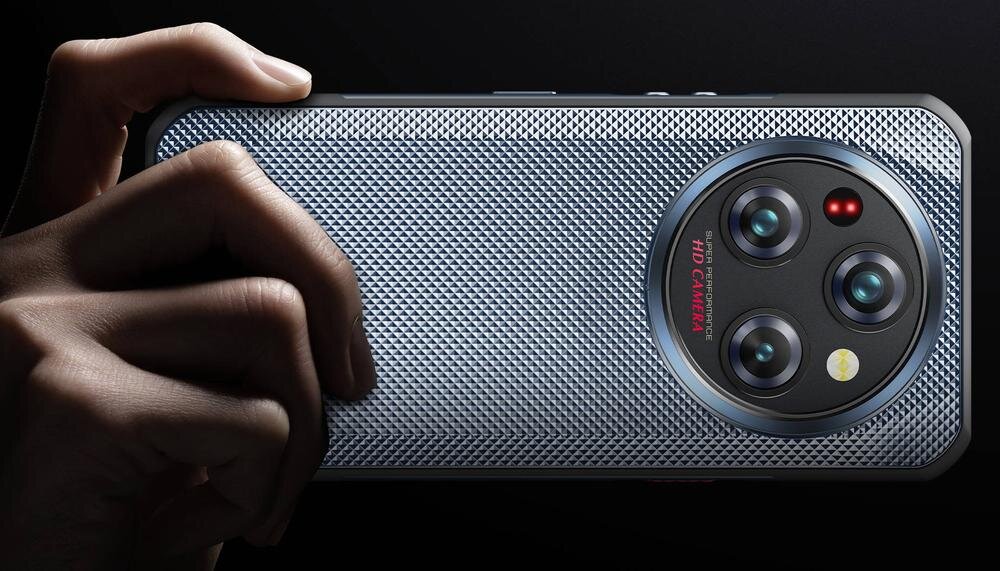 Osoba trzyma smartfon z teksturowaną obudową i trzema obiektywami aparatu. Na obudowie widoczny jest napis \'AI CAMERA\'.