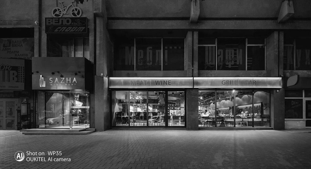 Czarno-białe zdjęcie przedstawia fasadę budynku z widocznymi szyldami \'SAZHA\' oraz \'MEAT WINE GRILL BAR\'. Na dole zdjęcia znajduje się napis \'Shot on WP35 OUKITEL AI camera\'.