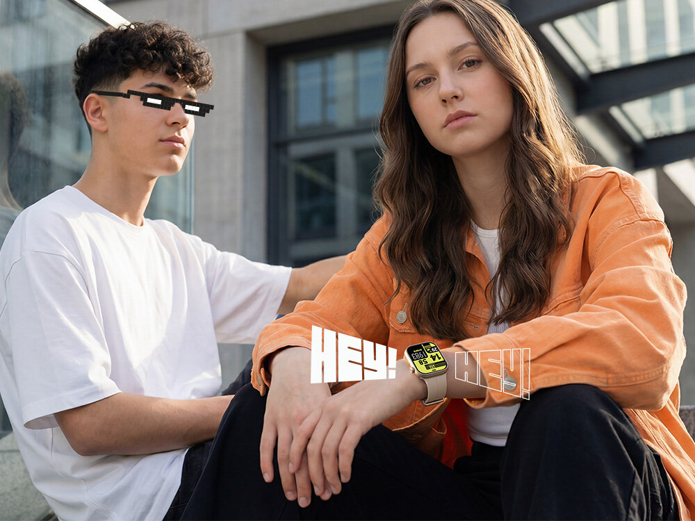 Dwie osoby siedzą na zewnątrz, jedna z nich nosi pomarańczową kurtkę i smartwatch na nadgarstku. Na obrazie widoczny jest tekst \'HEY!\' oraz ekran smartwatcha z wyraźnym wyświetlaczem.