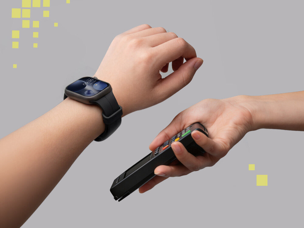 Osoba nosi czarny smartwatch na nadgarstku, zbliżając go do terminala płatniczego trzymanego przez inną osobę. Tło jest szare z żółtymi kwadratami w rogu.