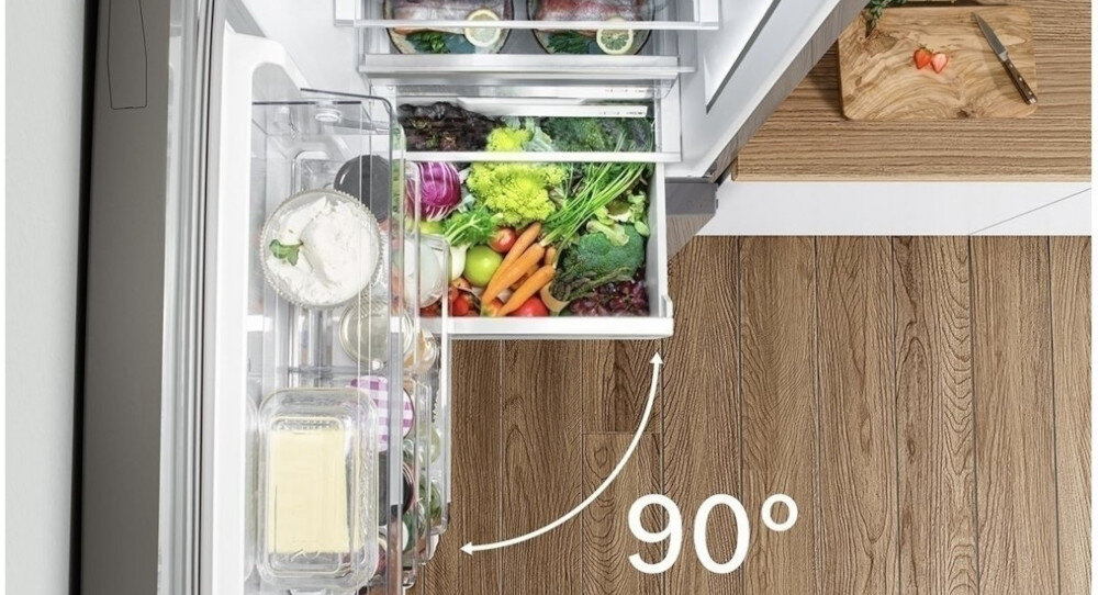 Otwarta lodówka z widocznymi szufladami wypełnionymi warzywami i innymi produktami spożywczymi. Na podłodze widoczny jest napis \'90°\'.