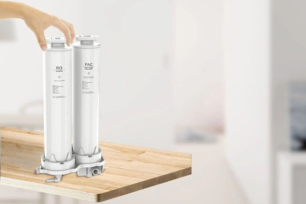 Na drewnianym blacie znajdują się dwa białe cylindryczne filtry z napisami \'RO\' i \'PAC\'. Widać rękę trzymającą jeden z filtrów.