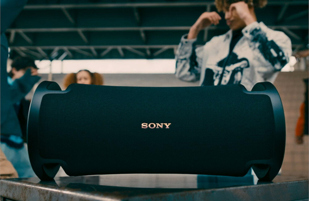 Czarny głośnik mobilny z widocznym logo \'SONY\' stoi na metalowej powierzchni. W tle znajdują się osoby, ale są one rozmyte i nieostre.