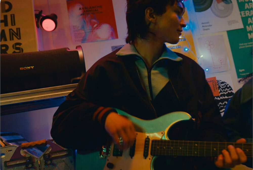 Osoba gra na gitarze elektrycznej w pomieszczeniu z plakatami na ścianie. W tle widoczny jest głośnik z logo \'SONY\'.