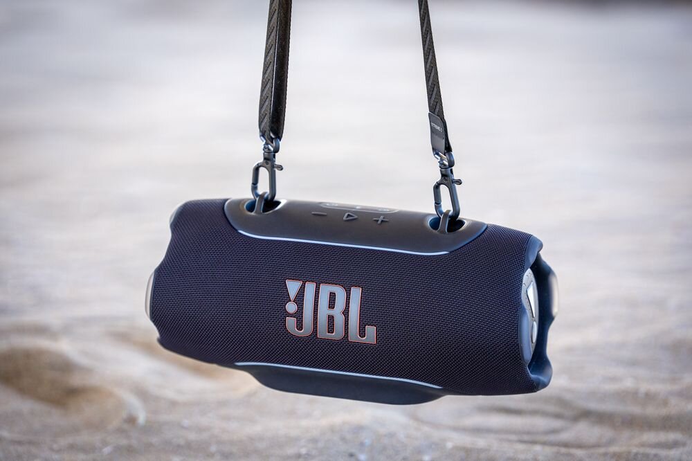 Czarny głośnik mobilny z logo \'JBL\' zawieszony na pasku, umieszczony na piasku. Na górze widoczne są przyciski sterujące.