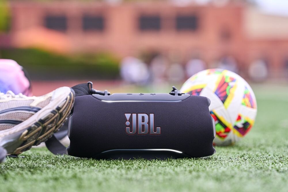 Na trawie znajduje się czarny głośnik z widocznym logo \'JBL\'. Obok leży kolorowa piłka i but sportowy.
