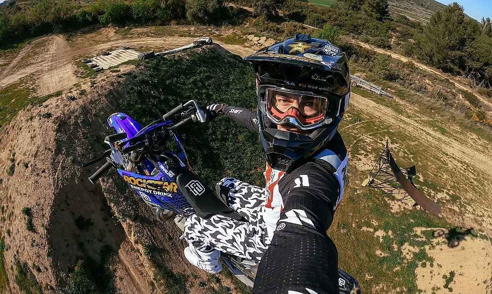 Osoba na motocyklu wykonuje akrobację w powietrzu na tle terenu z drzewami i rampą. Na motocyklu widoczne jest logo \'ROCKSTAR ENERGY DRINK\'.