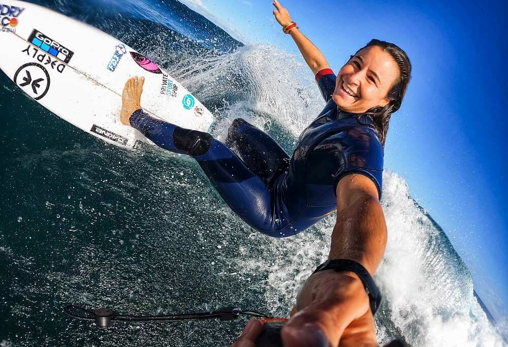 Osoba w kombinezonie surfingowym wykonuje manewr na fali, trzymając kamerę sportową. Na desce surfingowej widoczne są logotypy, w tym \'GOPRO\' i \'DARKY\'.