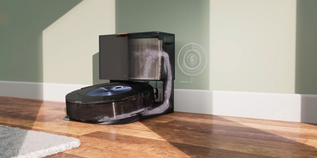 Robot sprzątający z czarną obudową znajduje się na drewnianej podłodze obok stacji dokującej przy ścianie. W tle widoczny jest schematyczny rysunek przedstawiający przepływ powietrza.
