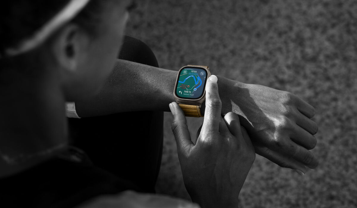 Osoba patrzy na smartwatch na nadgarstku, który wyświetla kolorowy ekran z aplikacją do śledzenia aktywności fizycznej. Pasek zegarka jest metalowy i ma jasny kolor.