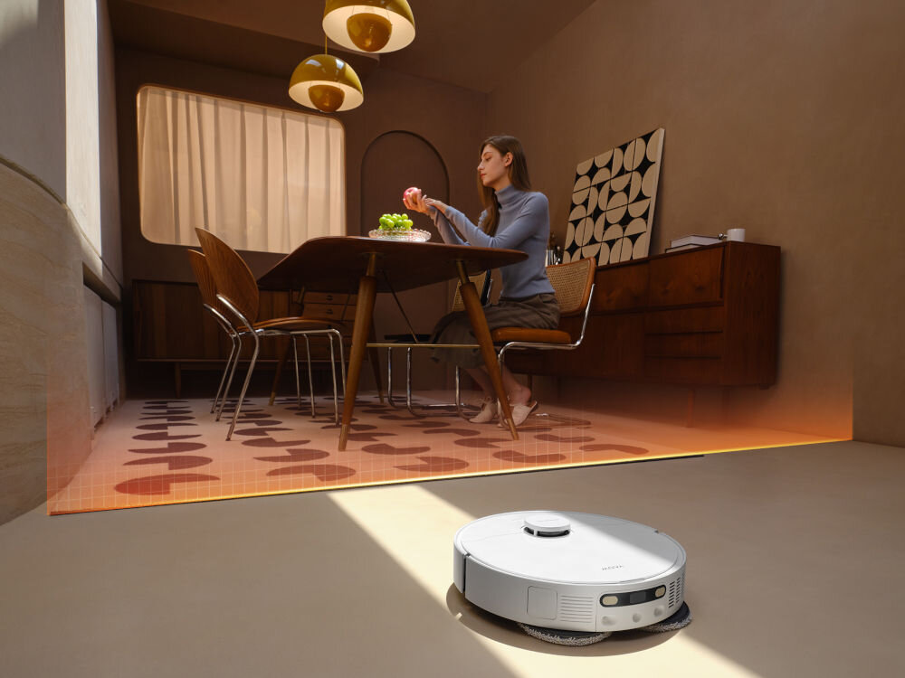 W jasnym pomieszczeniu znajduje się robot sprzątający na podłodze oraz osoba siedząca przy stole z owocami. W tle widać drewniane meble i dekorację ścienną.