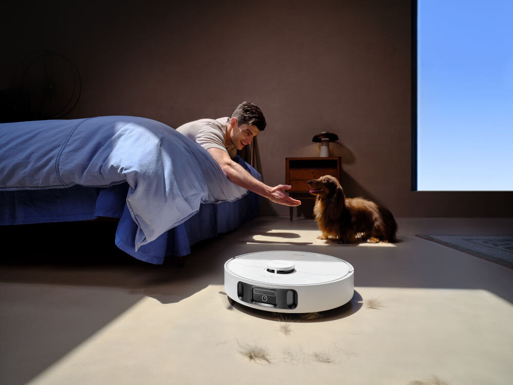 Na podłodze w pokoju znajduje się biały robot sprzątający, a obok niego leży kilka kępek sierści. W tle widać osobę siedzącą na łóżku i psa stojącego na podłodze.
