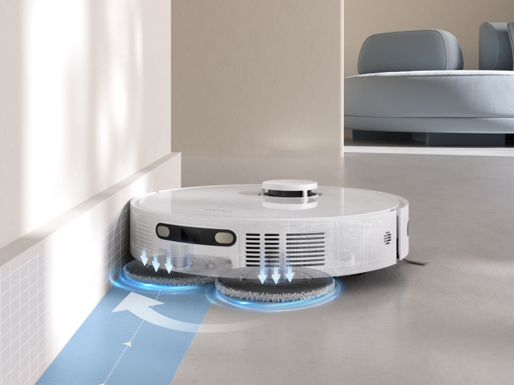Na obrazie widoczny jest biały robot sprzątający poruszający się wzdłuż ściany. Pod robotem znajdują się dwie okrągłe szczotki, a na podłodze widoczna jest niebieska strzałka wskazująca kierunek ruchu.