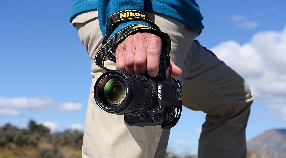Osoba trzyma aparat fotograficzny z widocznym logo Nikon, na tle nieba i krajobrazu. Aparat jest wyposażony w obiektyw z widocznymi oznaczeniami na pierścieniu.