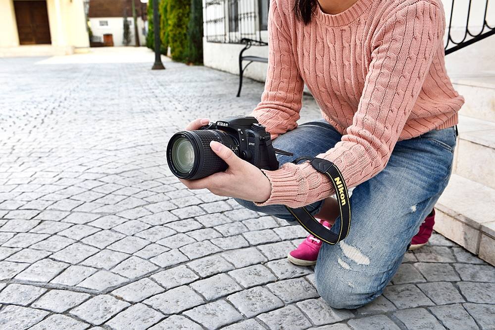 Osoba w różowym swetrze i dżinsach trzyma aparat fotograficzny z logo \'Nikon\' na pasku. Tło stanowi brukowana powierzchnia i fragment budynku.
