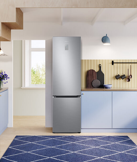 rb38c675cs9 elegancki design W kuchni znajduje się lodówka ze szczotkowanej stali nierdzewnej, obok której są niebieskie szafki i drewniane deski do krojenia na blacie. Na podłodze leży niebieski dywan z białym wzorem w romby.