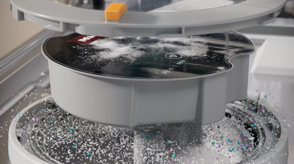 Obraz przedstawia wnętrze zmywarki z widocznym okrągłym pojemnikiem na detergent, na którym znajduje się logo Miele. Wokół pojemnika rozsypane są drobinki detergentu.