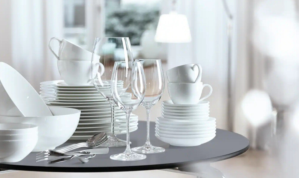 Na stole znajduje się zestaw białej porcelany, w tym talerze, miski, filiżanki oraz kieliszki do wina. W tle widoczne są rozmyte elementy wnętrza, takie jak lampa i okno.