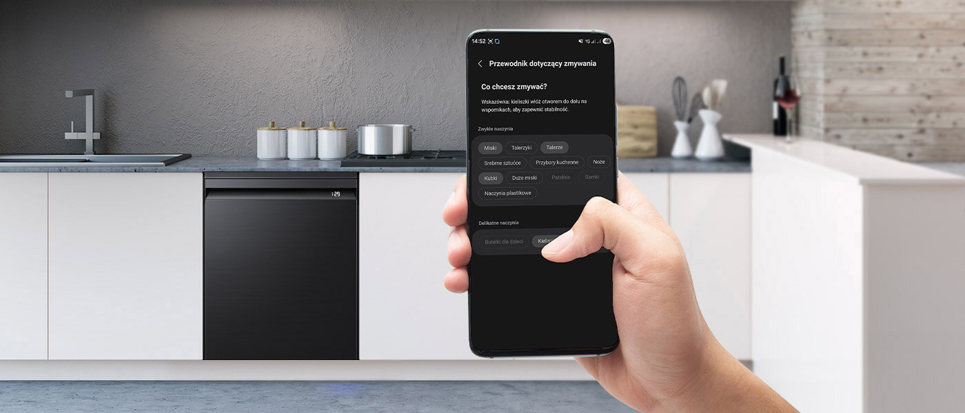 bez trudu efektywnie ułożysz naczynia w zmywarce Kuchnia z białymi szafkami i czarną zmywarką w zabudowie. Osoba trzyma smartfon z aplikacją dotyczącą zmywania, na ekranie widoczny jest tekst: \'Przewodnik dotyczący zmywania\'.