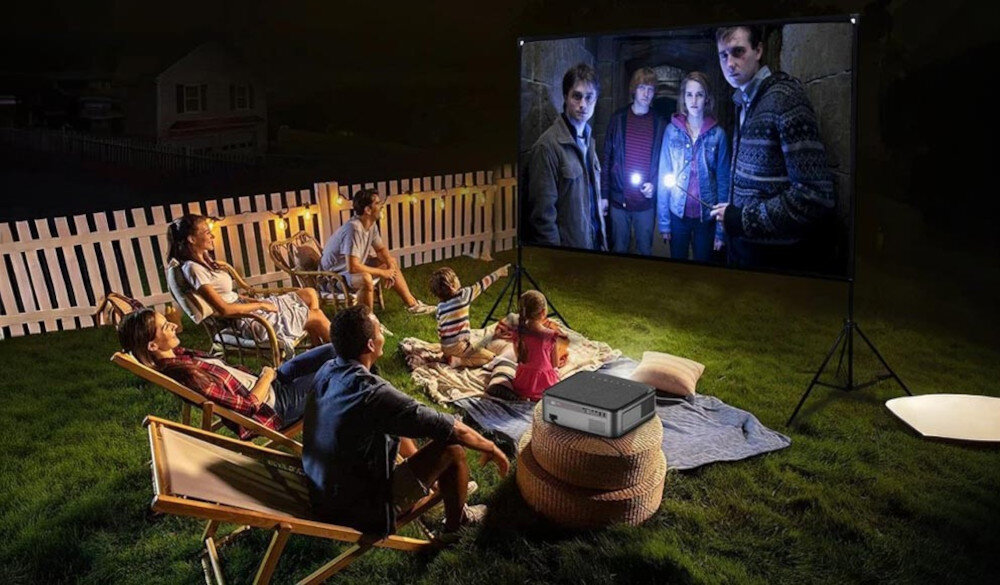Grupa osób siedzi na trawie przed ekranem projekcyjnym, na którym wyświetlany jest film. Obok ekranu znajduje się projektor, a w tle widoczny jest biały płot.