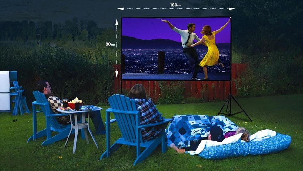 Na trawniku ustawiony jest ekran projekcyjny o wymiarach 160x90 cm, na którym wyświetlany jest film z tańczącą parą. Przed ekranem znajdują się osoby siedzące na niebieskich krzesłach i leżące na kocu.
