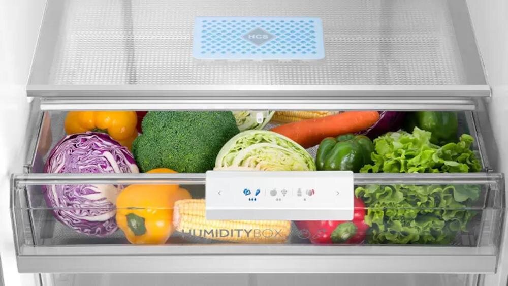 Wnętrze szuflady lodówki z różnorodnymi warzywami, takimi jak papryka, brokuł, kapusta i sałata. Na szufladzie widoczny napis \'HUMIDITY BOX\'.