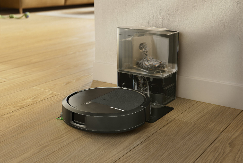 Czarny robot sprzątający znajduje się na drewnianej podłodze obok stacji dokującej z przezroczystym pojemnikiem na kurz. Na robocie widoczne jest logo \'iRobot\'.
