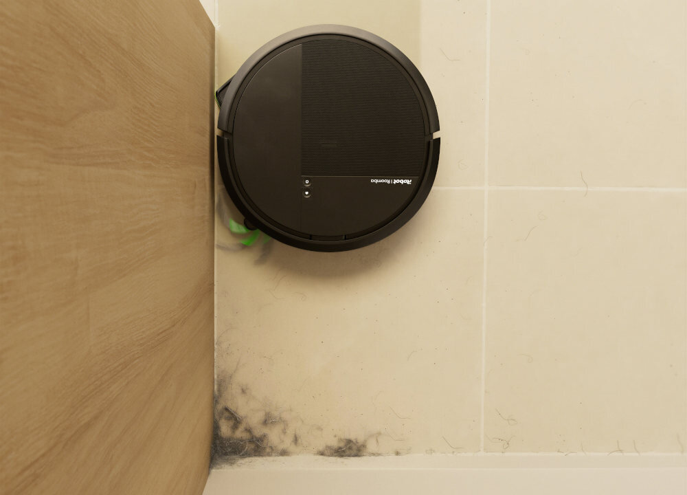 Czarny robot sprzątający znajduje się na jasnej podłodze przy drewnianej ścianie. Na robocie widoczne jest logo \'iRobot Roomba\'.