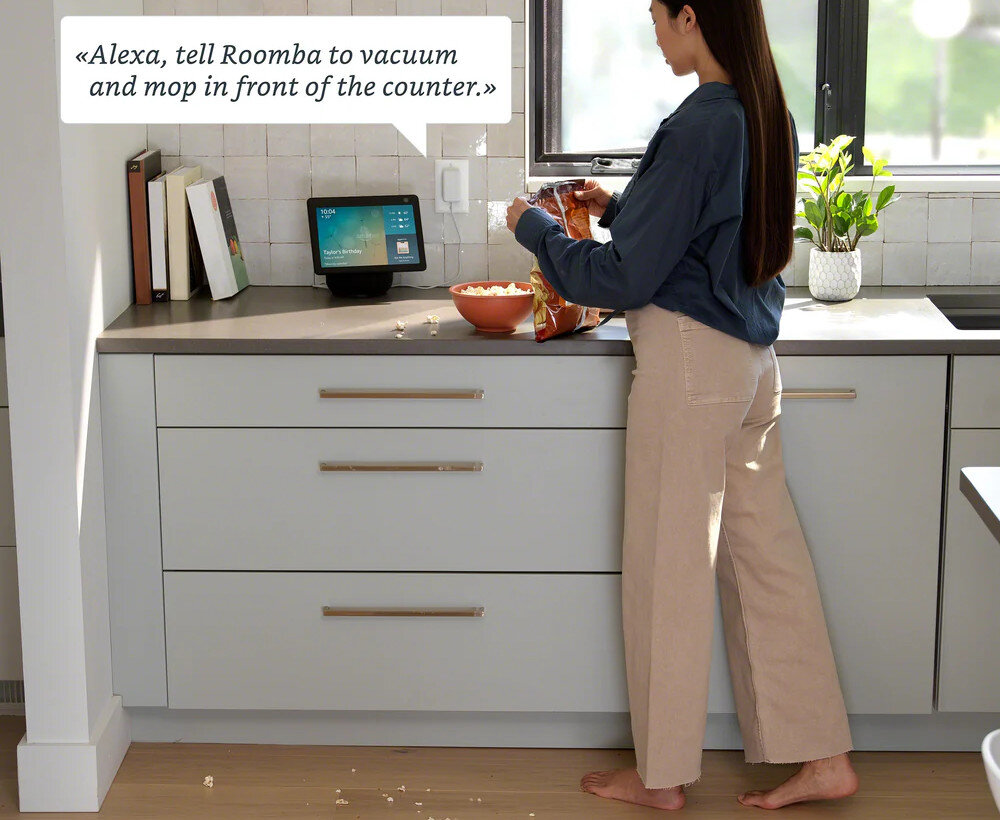 Kobieta stoi przy blacie kuchennym, trzymając otwartą paczkę chipsów. Na blacie znajduje się urządzenie z ekranem, na którym widoczny jest tekst: \'Alexa, tell Roomba to vacuum and mop in front of the counter.\'