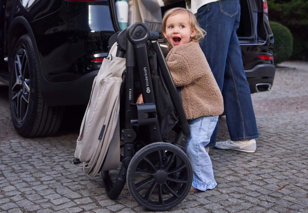 Złożony wózek dziecięcy KINDERKRAFT Vesto 2 Beżowy stoi obok samochodu. Obok wózka znajduje się dziecko w brązowej kurtce i dżinsach.