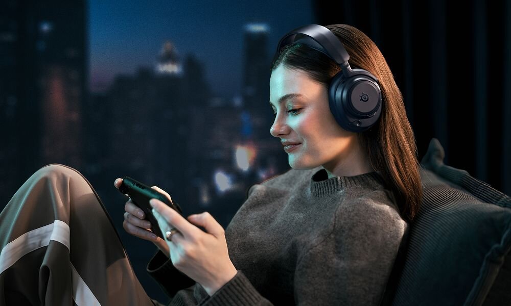 Osoba siedzi na kanapie, nosząc niebieskie słuchawki nauszne i patrzy na smartfon. W tle widoczne jest rozmyte miasto nocą.