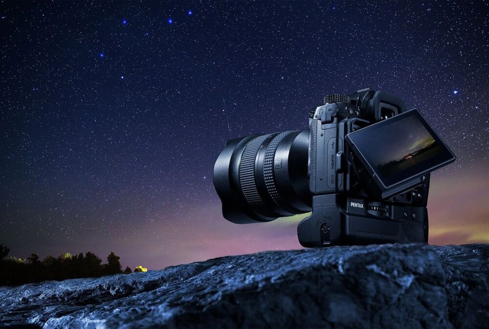 Na tle nocnego nieba z widocznymi gwiazdami znajduje się aparat fotograficzny z wysuniętym ekranem LCD. Na aparacie widoczny jest napis \'PENTAX\'.
