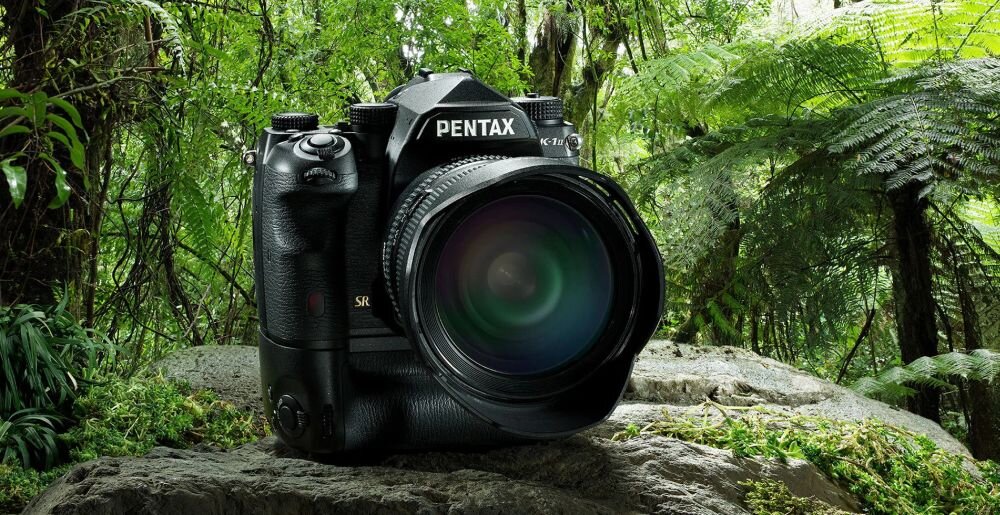 Aparat fotograficzny z logo \'PENTAX K-1\' umieszczony na kamieniu w otoczeniu zielonej roślinności.