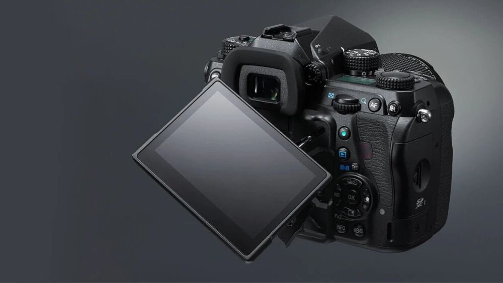 Na obrazie widoczny jest aparat fotograficzny z odchylanym ekranem LCD. Aparat ma czarną obudowę z widocznymi przyciskami i pokrętłami sterującymi.