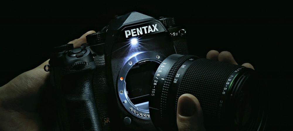 Na ciemnym tle widoczny jest aparat fotograficzny z logo PENTAX, z odłączonym obiektywem trzymanym w dłoni.