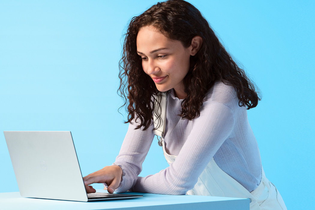Osoba korzysta z laptopa na tle jasnoniebieskiej ściany. Laptop jest otwarty i ma srebrną obudowę.