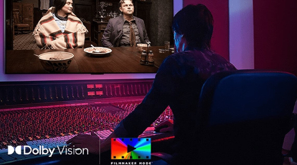 Osoba siedzi przed dużym ekranem, na którym wyświetlana jest scena z dwoma osobami przy stole. Na dole ekranu widoczne są loga \'Dolby Vision\' i \'Filmmaker Mode\'.