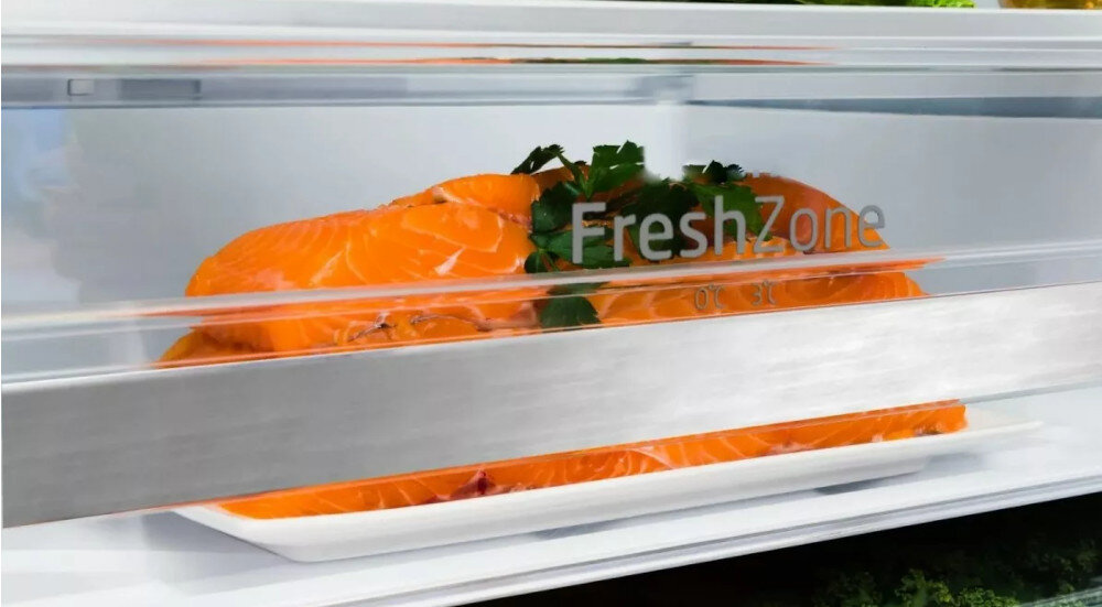 Wnętrze lodówki z przezroczystą szufladą oznaczoną napisem \'FreshZone\', w której znajduje się kawałek łososia udekorowany zielonymi liśćmi.