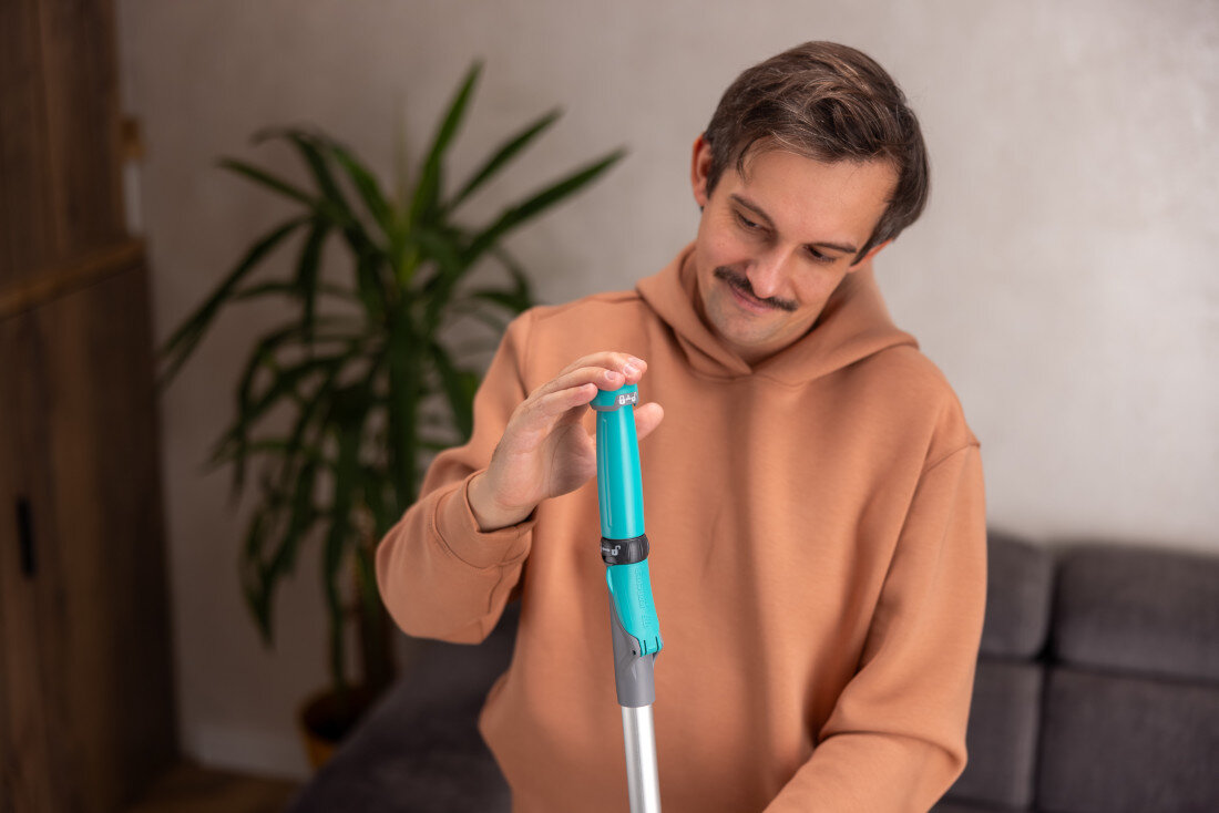 Osoba trzyma mop z turkusowym uchwytem w pomieszczeniu z rośliną i sofą w tle.