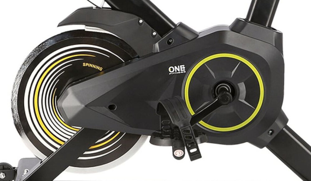 Część roweru spinningowego z widocznym mechanizmem napędowym i pedałem. Na obudowie znajduje się logo \'ONE FITNESS\' oraz napis \'SPINNING\'.