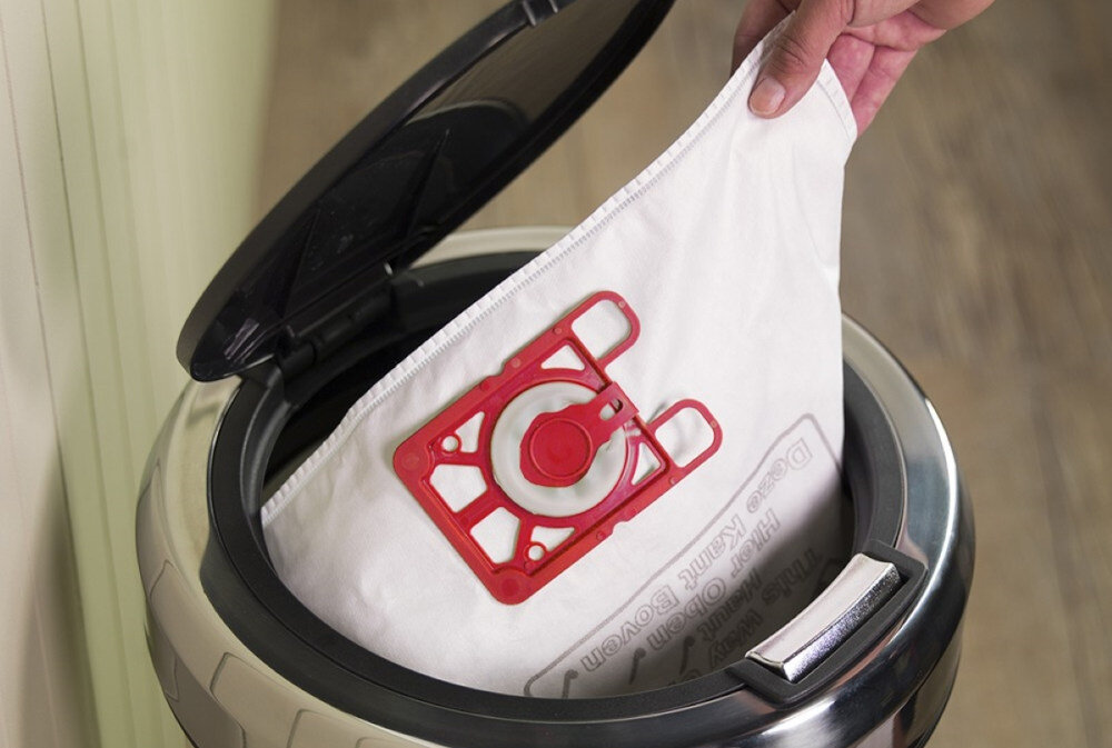 Osoba wkłada worek do odkurzacza z otwartą pokrywą. Worek ma czerwony plastikowy element mocujący.