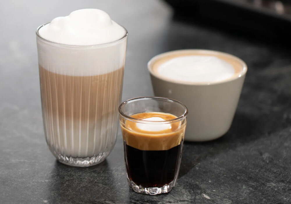 Na blacie znajdują się trzy różne napoje kawowe: wysoka szklanka z warstwami kawy i mleka, mała filiżanka z kawą z pianką oraz szklanka z espresso z kremową pianką na wierzchu.