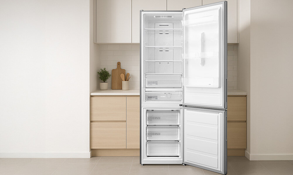 Otwarta lodówka w kolorze inox z widocznymi półkami i szufladami, ustawiona w nowoczesnej kuchni z jasnymi szafkami. W tle znajduje się blat z rośliną i deską do krojenia.