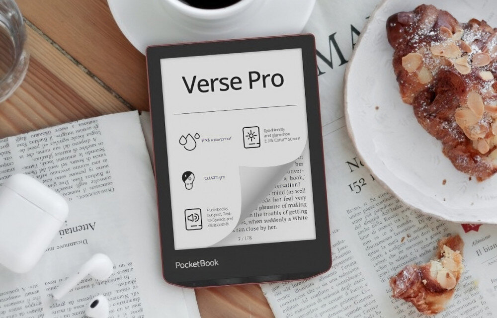 Na drewnianym stole leży czytnik e-booków z widocznym napisem \'Verse Pro\' na ekranie. Obok znajdują się gazeta, filiżanka kawy, talerz z ciastkiem oraz słuchawki bezprzewodowe.