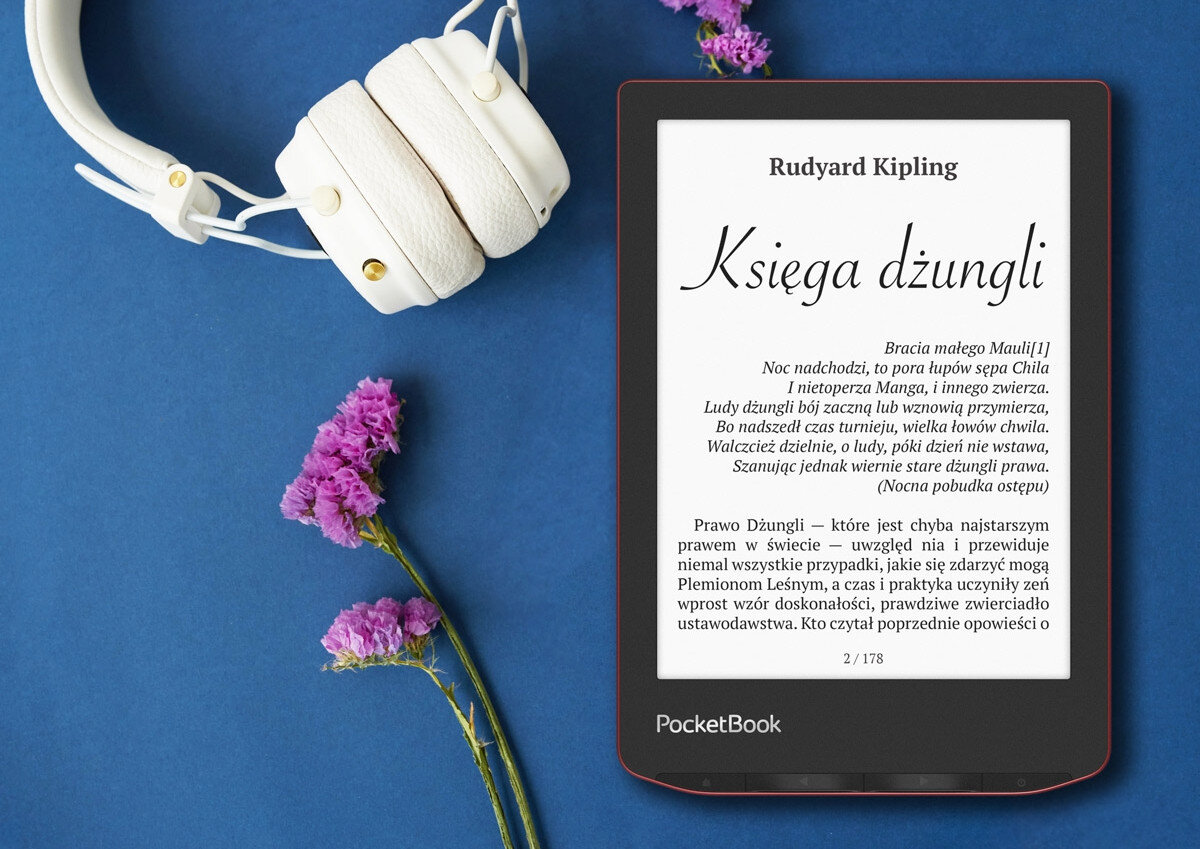 Na niebieskim tle leży czerwony czytnik e-booków z wyświetlonym tekstem \'Rudyard Kipling Księga dżungli\' oraz białe słuchawki i fioletowe kwiaty. Na dole ekranu czytnika widnieje napis \'PocketBook\'.