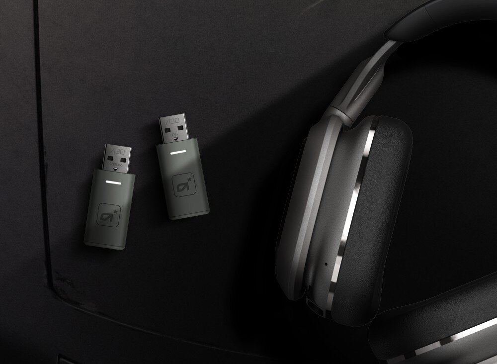 Na ciemnym tle znajdują się dwa adaptery USB z logo oraz bezprzewodowe słuchawki nauszne w kolorze srebrno-czarnym.