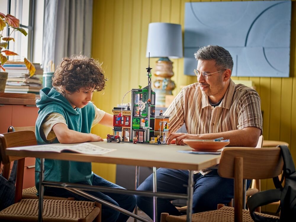 Dwie osoby siedzą przy stole, na którym znajduje się zestaw klocków LEGO przedstawiający budynek. W tle widać lampę i obraz na ścianie.