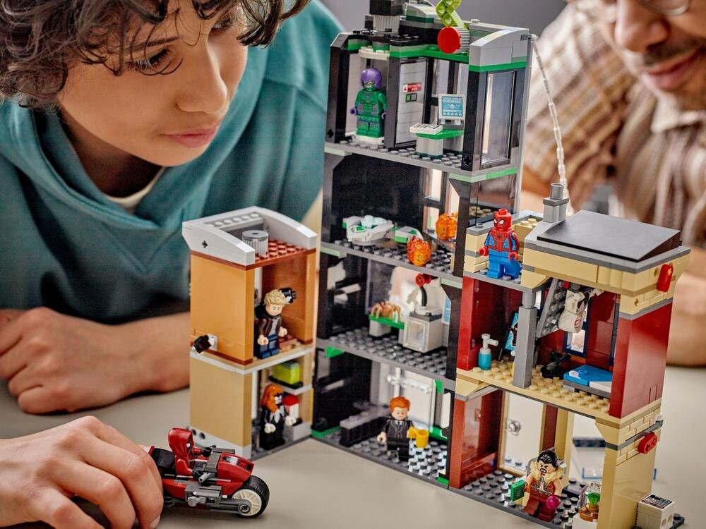 Dwoje dzieci bawi się zestawem klocków LEGO przedstawiającym wielopiętrowy budynek z postaciami superbohaterów. Na pierwszym planie widoczny jest czerwony motocykl z klocków.