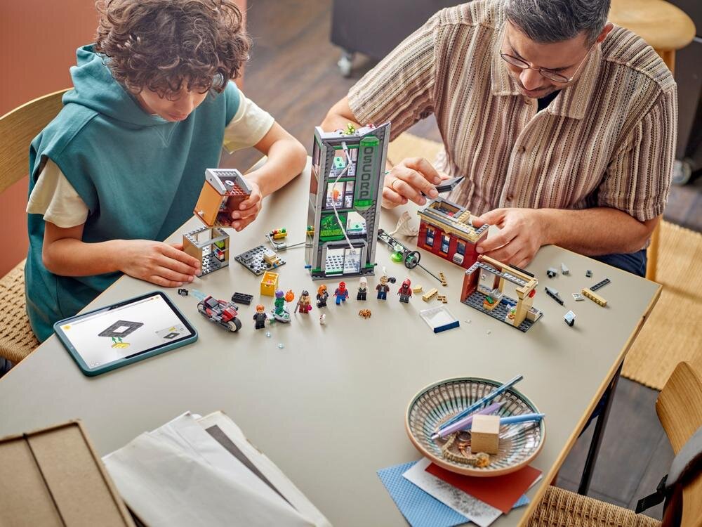 Na stole znajduje się zestaw klocków LEGO z budynkiem i figurkami, a dwie osoby składają elementy. Widoczny jest napis \'OSCORP\' na jednym z elementów budynku.